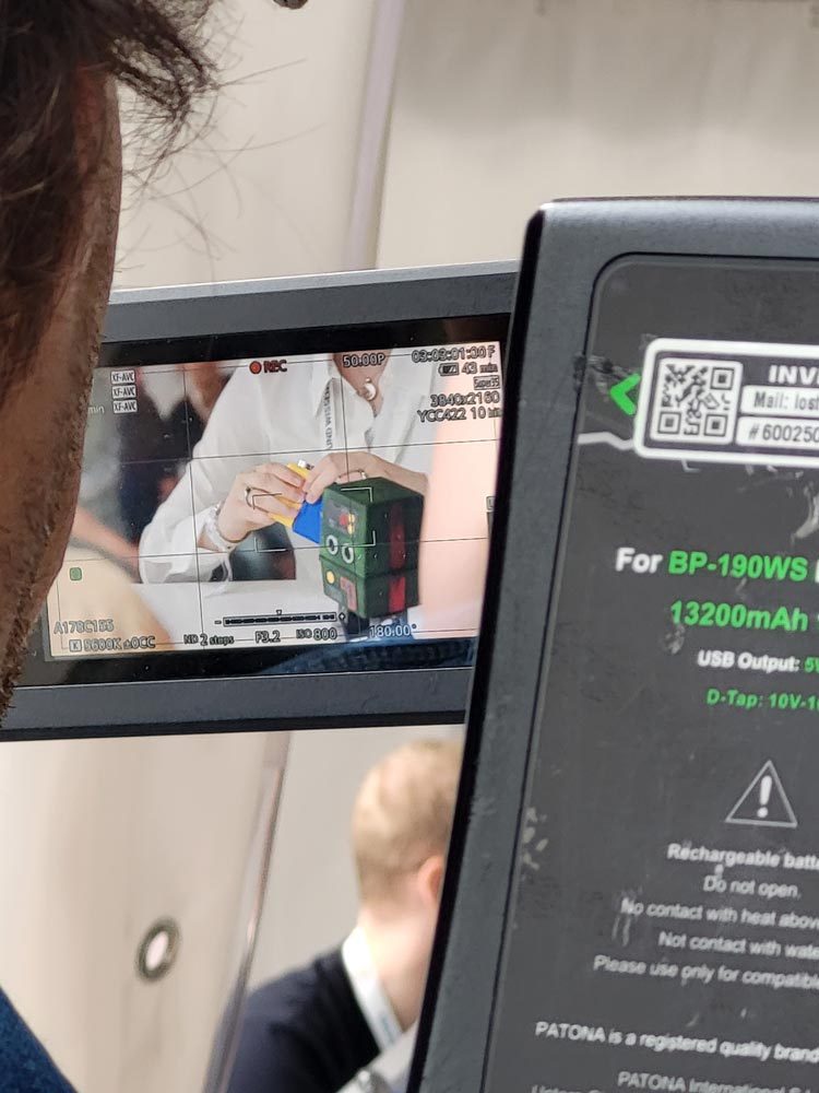 Eine Person hält ein kleines quadratisches Objekt mit Knöpfen und Wählscheiben. Auf dem Bildschirm einer Digitalkamera ist die Szene zu sehen, verschiedene Bedieneinstellungen sind sichtbar.