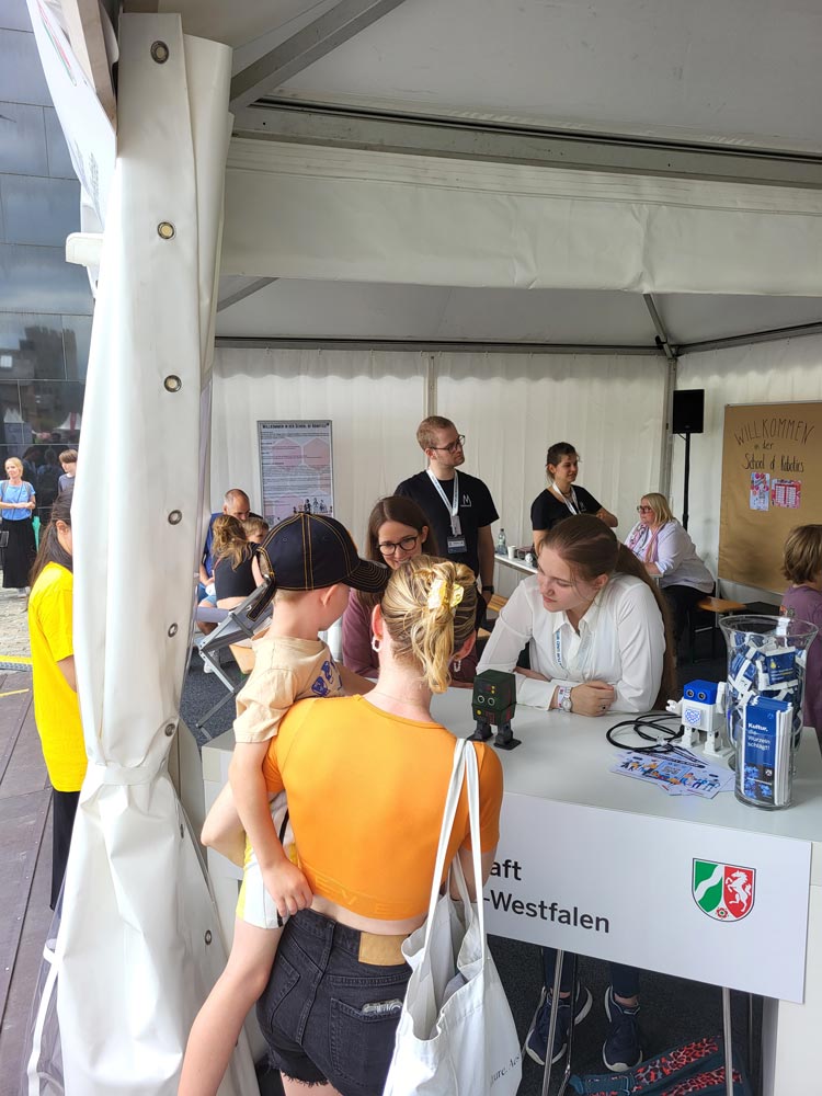 Menschen unterhalten sich an einem Stand in einem weißen Zelt bei einer Veranstaltung. Eine Frau und ein Kind, von hinten zu sehen, unterhalten sich mit Mitarbeitern hinter einem Tisch mit der Aufschrift „Westfalen“, auf dem verschiedene Gegenstände und ein Schild ausgestellt sind. Die lebhafte Szene ist Teil der Feierlichkeiten zum NRW-Tag 2024.