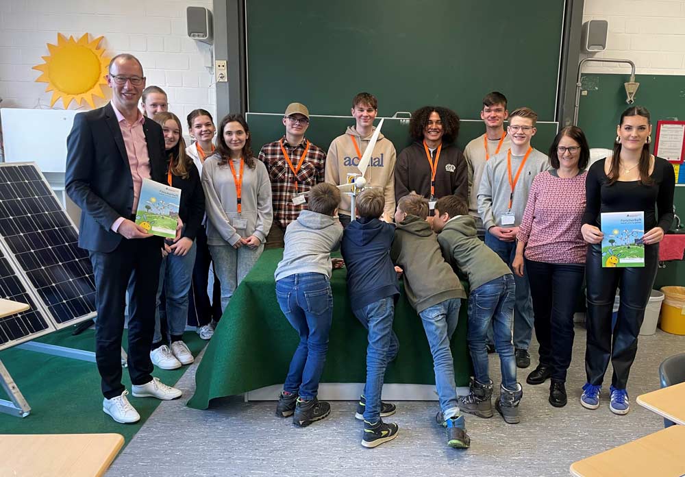 Schüler und Lehrer sind in einem Klassenzimmer versammelt und stehen um ein Display mit einem Solarpanel herum. Sie halten Bücher in den Händen und lächeln. Im Hintergrund hängt eine Sonnendekoration an der Wand.