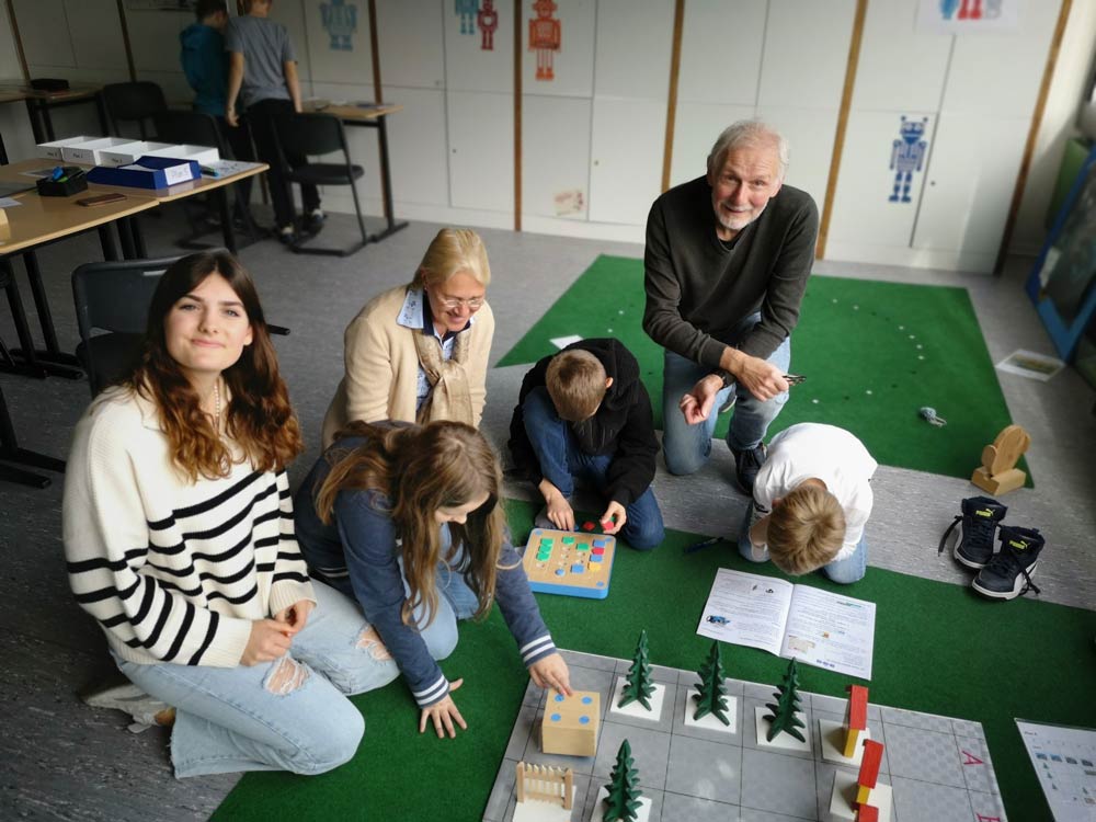 Die Leute sitzen um einen Tisch herum und spielen in einem Klassenzimmer pädagogische Brettspiele. Links im Bild sitzt Mara Meier in einem gestreiften Pulli und lächelt in die Kamera.