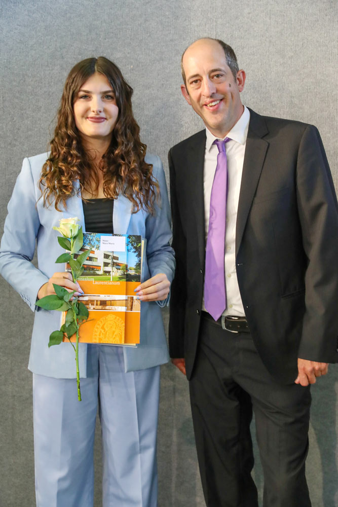 Eine junge Frau (Mara Maier) in einem hellblauen Anzug hält eine Rose und eine Urkunde in den Händen und steht neben einem Mann in einem dunklen Anzug und einer violetten Krawatte. Sie stehen vor einem grauen Hintergrund.