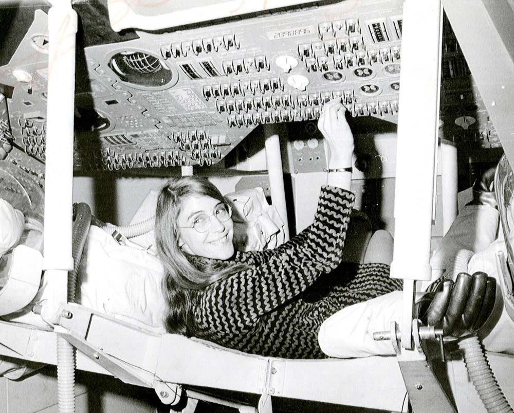 Das Bild ist eine schwarz-weiß Fotografie. Eine Frau (Margaret Hamilton) liegt in einem Astronautensitz, blickt in die Kamera und bedient Schalter an einem Bedienfeld an der Decke.
