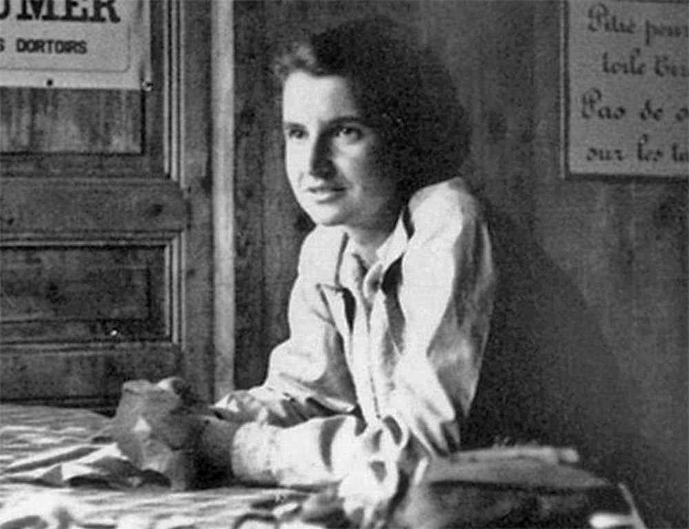 Das Bild ist eine schwarz-weiß Fotografie. Eine Frau (Rosalind Franklin) lehnt sich an einem Tisch nach vorne und legt ihre Hände auf die Tischplatte. Im Hintergrund sind Holzpaneele und teilweise sichtbare Schilder zu sehen.