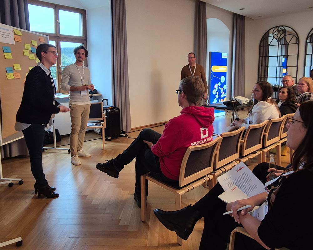 Eine Gruppe von Menschen nimmt an einer Workshop-Sitzung der Veranstaltung „Zukunftsberufe für Mädchen“ teil, die im Rahmen des „zdi-Heldinnen-Oktober“ stattfindet. Eine Person präsentiert vor einer mit Haftnotizen bedeckten Tafel. Die anderen sitzen da und hören aufmerksam zu.