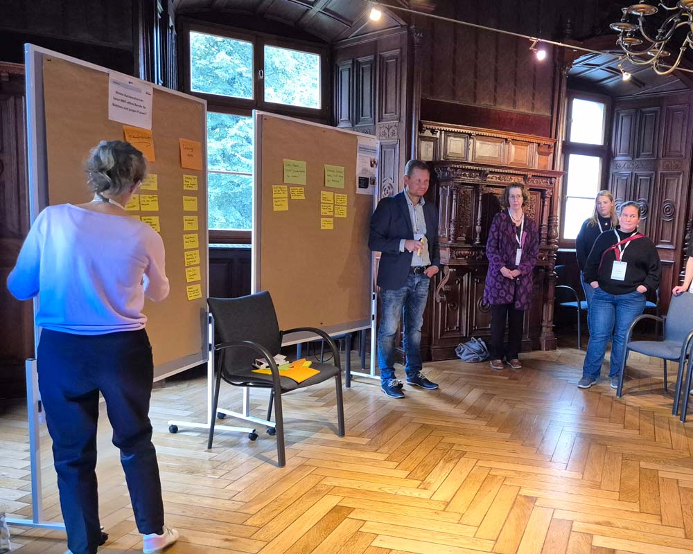 In einem holzgetäfelten Raum diskutieren Menschen über Ideen. Zwei große Tafeln mit Notizen sind zu sehen, im Vordergrund stehen zwei leere Stühle. Die Szene ist Teil der Veranstaltung „Zukunftsberufe für Mädchen“, die im Rahmen des „zdi-Heldinnen-Oktober“ stattfindet.