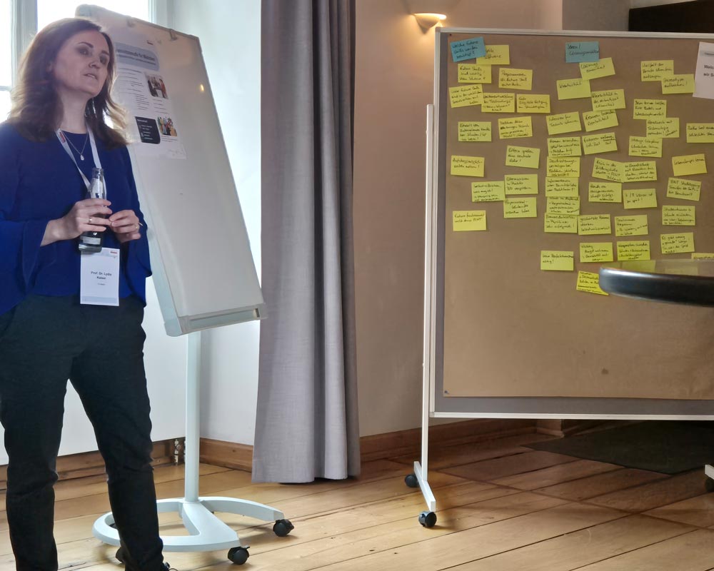 Prof. Dr. Lydia Kaiser steht neben einem Flipchart und einer mit Haftnotizen bedeckten Tafel in einem Raum. Sie hält eine Flasche und trägt ein Namensschild. Sie ist Teil der Veranstaltung „Zukunftsberufe für Mädchen“, die im Rahmen des „zdi-Heldinnen-Oktober“ stattfindet.