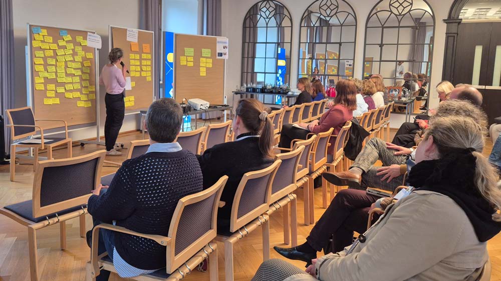 Bei der Abschlusspräsentation der Veranstaltung „Zukunftsberufe für Mädchen“, die im Rahmen des „zdi-Heldinnen-Oktober“ stattfindet, sitzt eine Gruppe von Menschen in einem Konferenzraum und hört einer Frau zu, die auf einem mit Haftnotizen bedeckten Whiteboard steht.