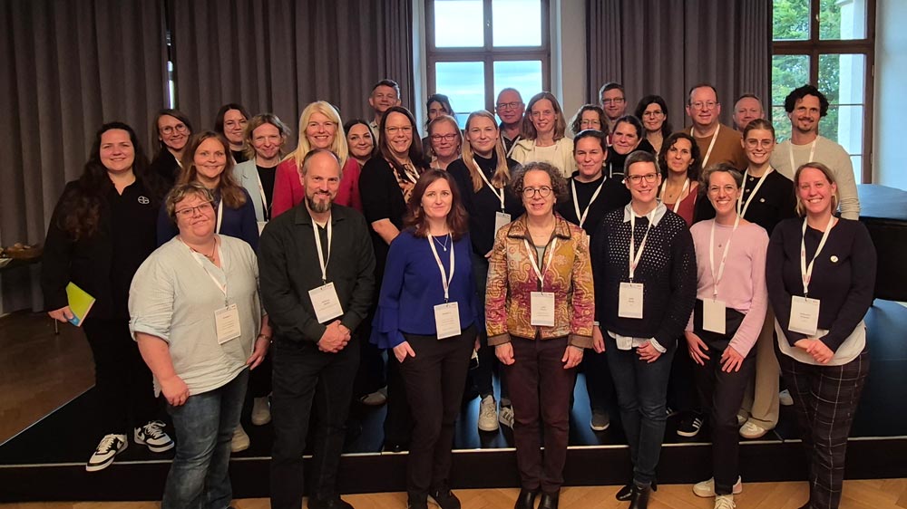 Eine Gruppe von Menschen steht in einem Innenbereich zusammen, trägt Schlüsselbänder und posiert für ein Gruppenfoto vor großen, mit Vorhängen verhangenen Fenstern. Das Foto zeigt die Teilnehmenden der Veranstaltung "Zukunftsberufe für Mädchen", die im Rahmen des zdi-Heldinnen-Oktobers stattfand.