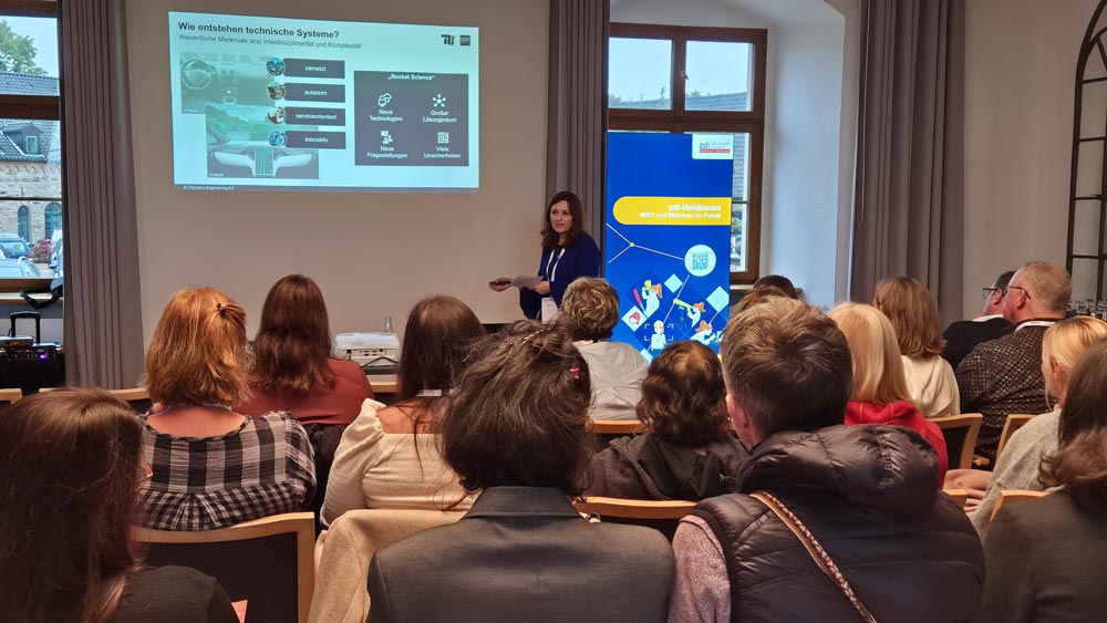 Prof. Dr, Lydia Kaiser steht vor einem sitzenden Publikum der Veranstaltung "Zukunftsberufe für Mädchen" und hält einen Vortrag. Auf einem Bildschirm wird eine Folie mit Text und Bildern auf Deutsch angezeigt. Im Hintergrund ist ein Banner zu sehen, das für „zdi-Heldinnen-Oktober“ wirbt vorstellt.