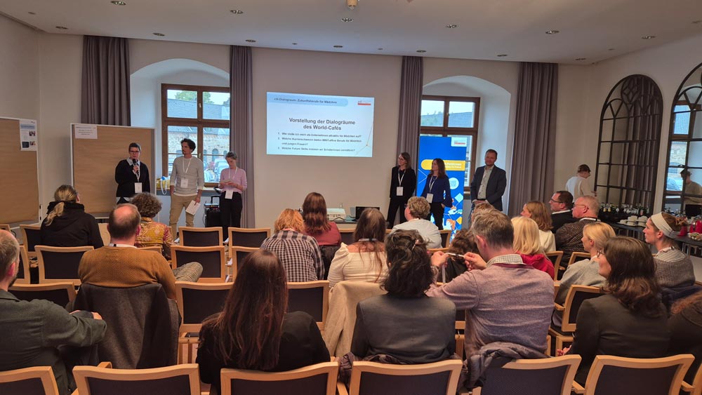 Eine Gruppe von Menschen in einem Konferenzraum hört sich einen Vortrag zum Thema „Zukunftsberufe für Mädchen“ an. Vorne stehen mehrere Vortragende, hinter ihnen läuft eine projizierte Präsentation. Diese Veranstaltung des zdi-Heldinnen-Oktobers fesselt die Anwesenden, die den Vortragenden gegenübersitzen.