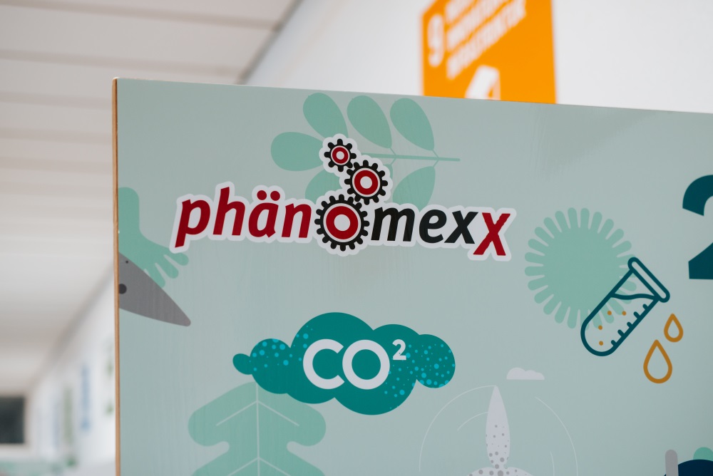 Das Bild zeigt ein Schild mit dem Text „PhänomexX Krefeld“, begleitet von Abbildungen von Zahnrädern, einem CO₂-Symbol, einem Reagenzglas und einem Tropfen auf hellem Hintergrund.