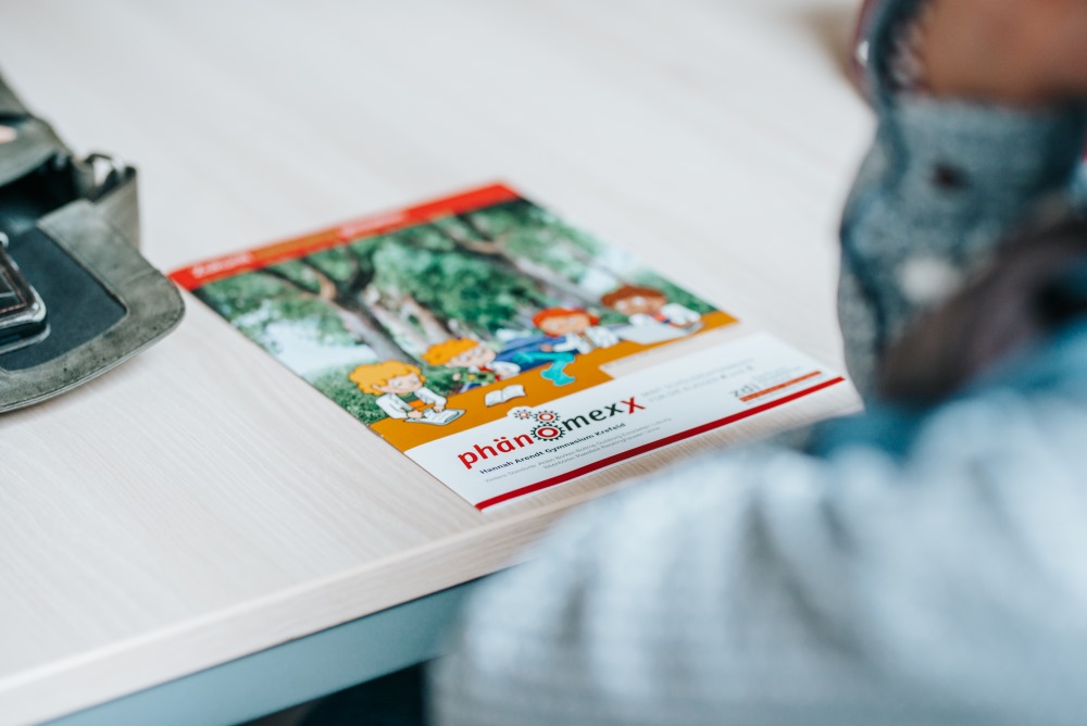 Eine Person betrachtet ein bunt illustriertes PhänomexX-Forscherheft auf dem Tisch, mit Comicfiguren, Bäumen und spannenden Informationen.