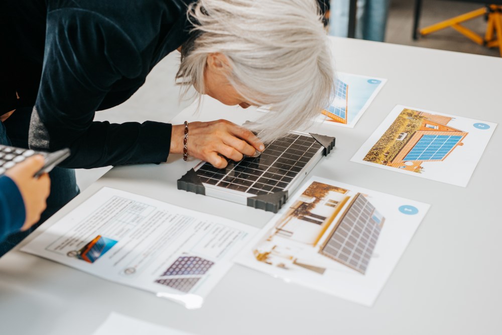 Eine Person untersucht auf einem Tisch im PhänomexX-Labor Krefeld das Modell eines Solarpanels. Der Tisch ist umgeben von Dokumenten und Fotos von Solaranlagen.