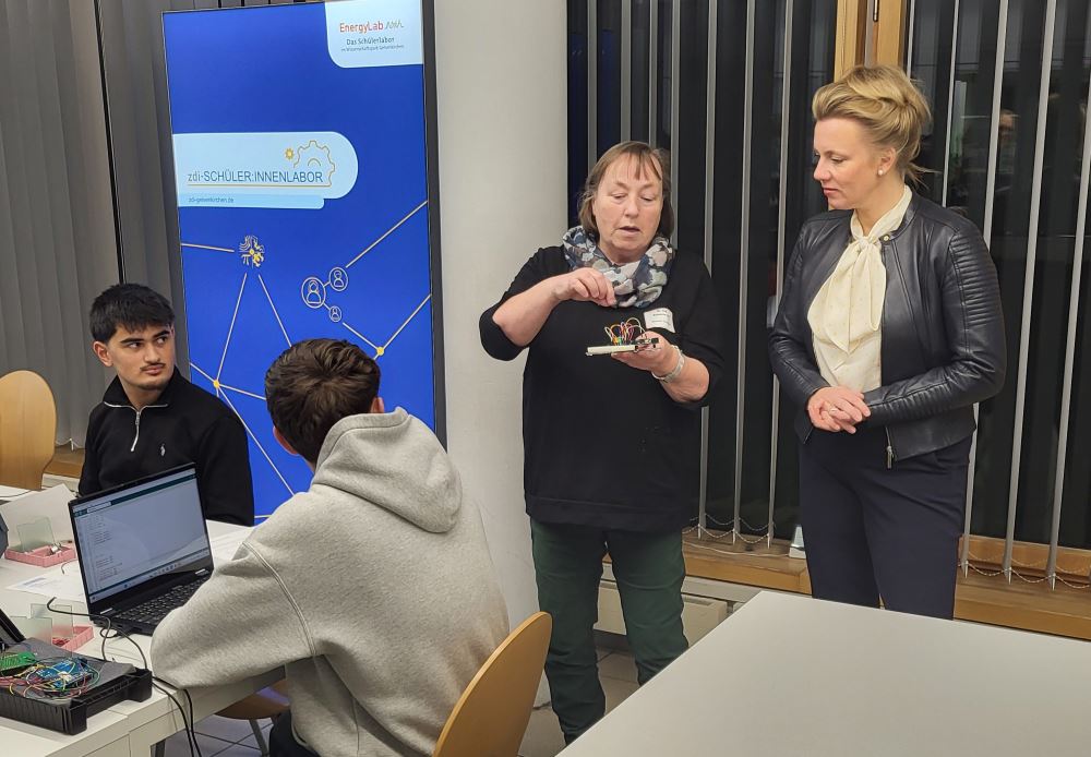 Zwei Personen diskutieren an einem Tisch über Schaltkreiskomponenten, während zwei andere zuschauen. Auf dem Tisch liegen Laptops und elektronische Geräte. Ein Schild und Fenster dienen als Hintergrund für diese kollaborative und innovative Szene.