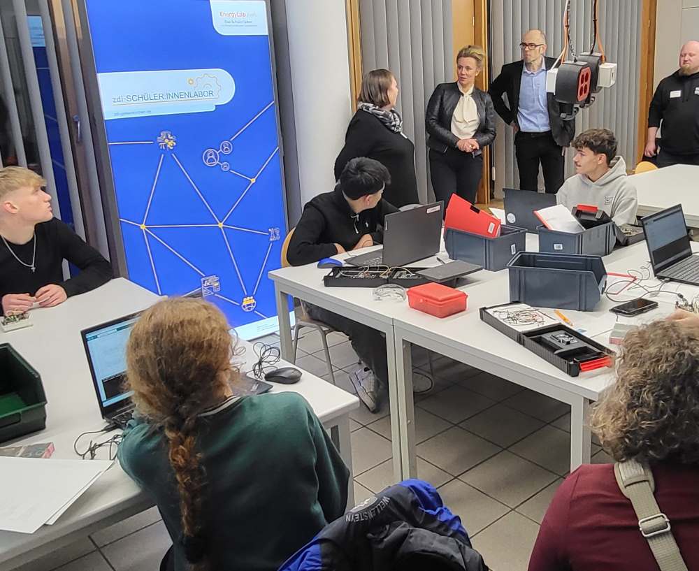 In einem belebten Raum versammeln sich Menschen um Computer und Geräte. Einige junge Leute sitzen an Schreibtischen, während andere vor einem blauen Bildschirm stehen.
