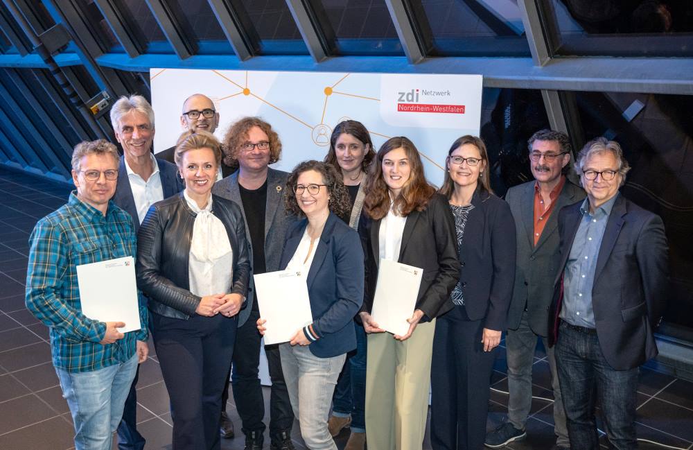 Eine Gruppe von Menschen steht drinnen mit Urkunden, darunter Förderbescheide, vor einem Transparent mit der Aufschrift „zdi Netzwerk Nordrhein-Westfalen“.