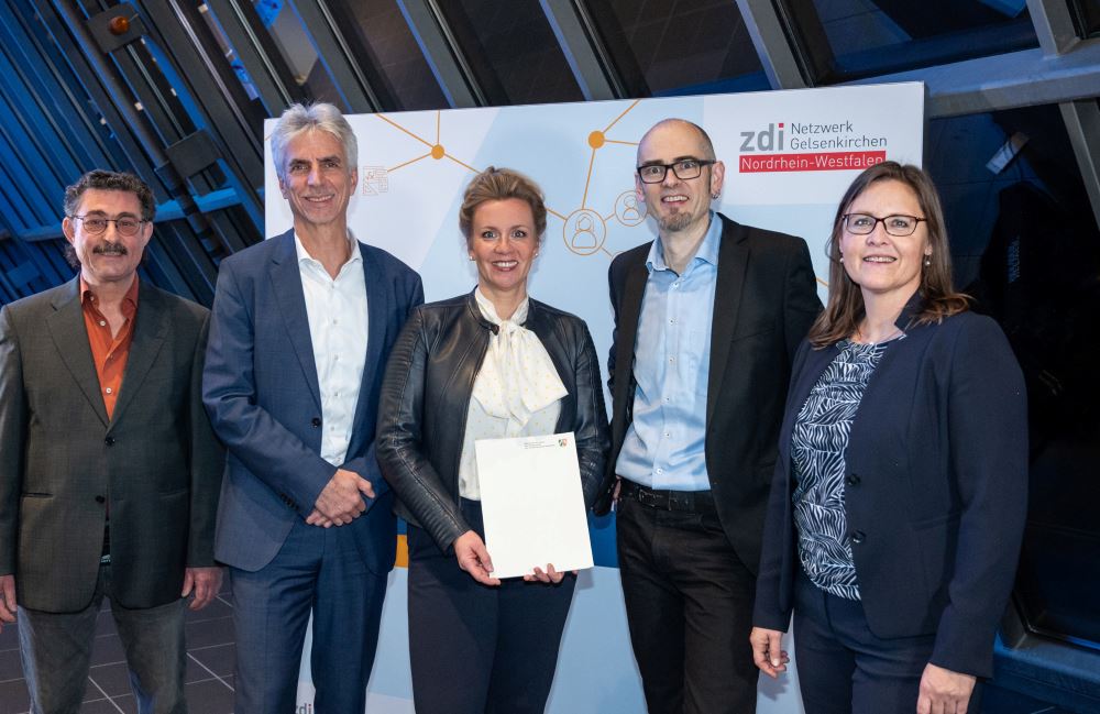 Fünf Personen stehen drinnen und lächeln in die Kamera. Eine Person hält stolz eine Förderbescheid-Urkunde in den Händen. Im Hintergrund sorgt ein Banner mit Logos für die festliche Atmosphäre.