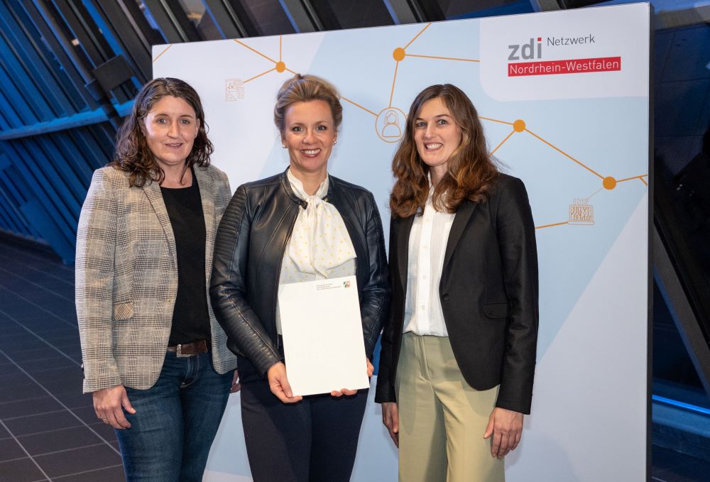Drei Frauen stehen vor einem Schild des zdi-Netzwerk Nordrhein-Westfalen und halten stolz einen Förderbescheide-Zettel in der Hand.