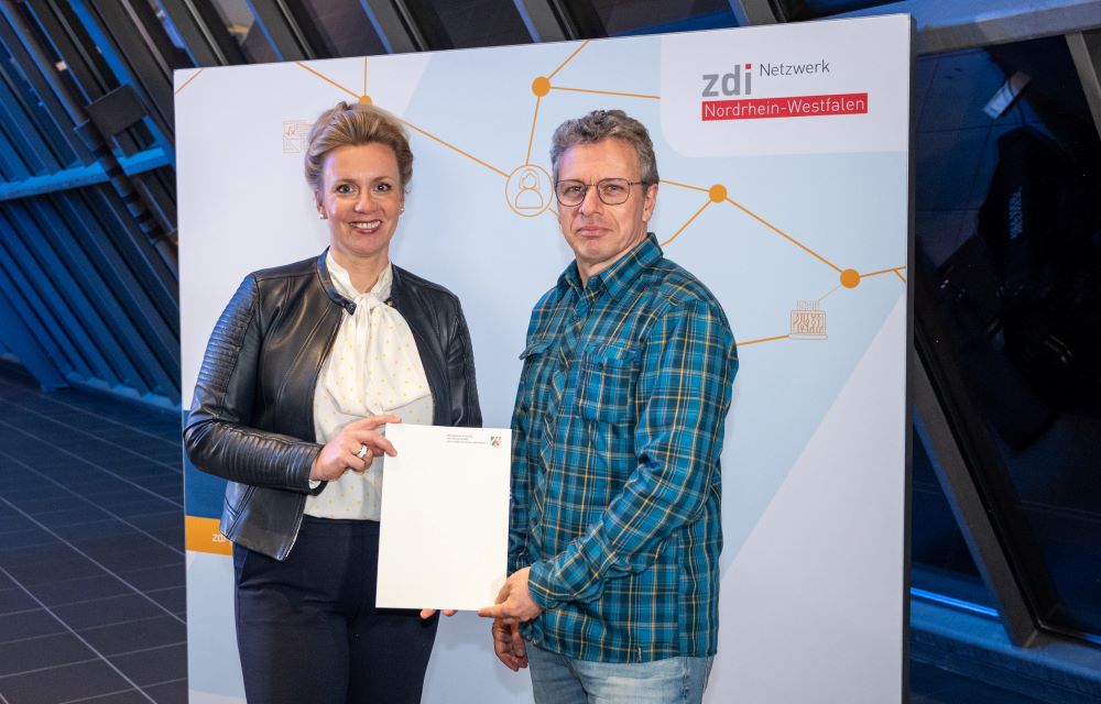 Zwei Personen stehen drinnen vor der Kulisse des zdi-Netzwerk Nordrhein-Westfalen, eine Person hält ein Förderbescheide-Dokument in der Hand.