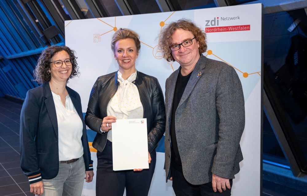 Drei Personen stehen drinnen neben einem Schild. Sie tragen legere Business-Kleidung und lächeln strahlend. In der Mitte hält eine Person stolz ein Dokument in der Hand, wahrscheinlich einen wichtigen Förderbescheid für ihr gemeinsames Projekt.
