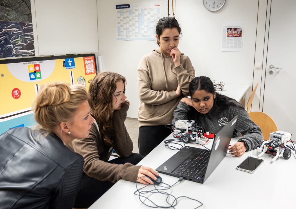 Vier Frauen sitzen um einen Tisch in einem Raum, der mit pädagogischen Postern dekoriert ist. Sie konzentrieren sich auf ein Projekt mit einem Laptop und Robotergeräten und zeigen damit ihr innovatives Streben nach Wissen.