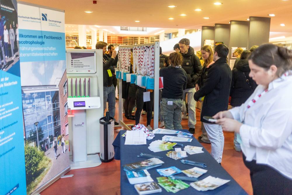 Bei der Veranstaltung KReateFUTURE 2025 erkunden die Teilnehmenden Broschüren auf einem Tisch, eine dynamische Posteranzeige betrachten und interagieren mit einem digitalen Kiosk.