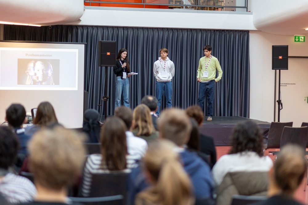 Drei Personen stehen auf einer Bühne bei KReateFUTURE 2025 und präsentieren einem interessierten Publikum ihre Ideen. Hinter ihnen ist auf einer großen Leinwand das Wort „Posthumanismus“ zu sehen, das den Ton für die Erforschung zukünftiger Möglichkeiten angibt.