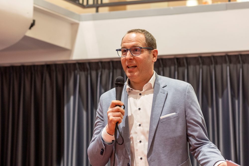 Ein Mann in einem hellblauen Anzug und mit Brille, Dr. Dominik Paul, steht im Innenbereich vor dunklen Vorhängen steht und hält ein Mikrofon. Er begrüßt die Teilnehmenden bei der Veranstaltung KReateFUTURE 2025.