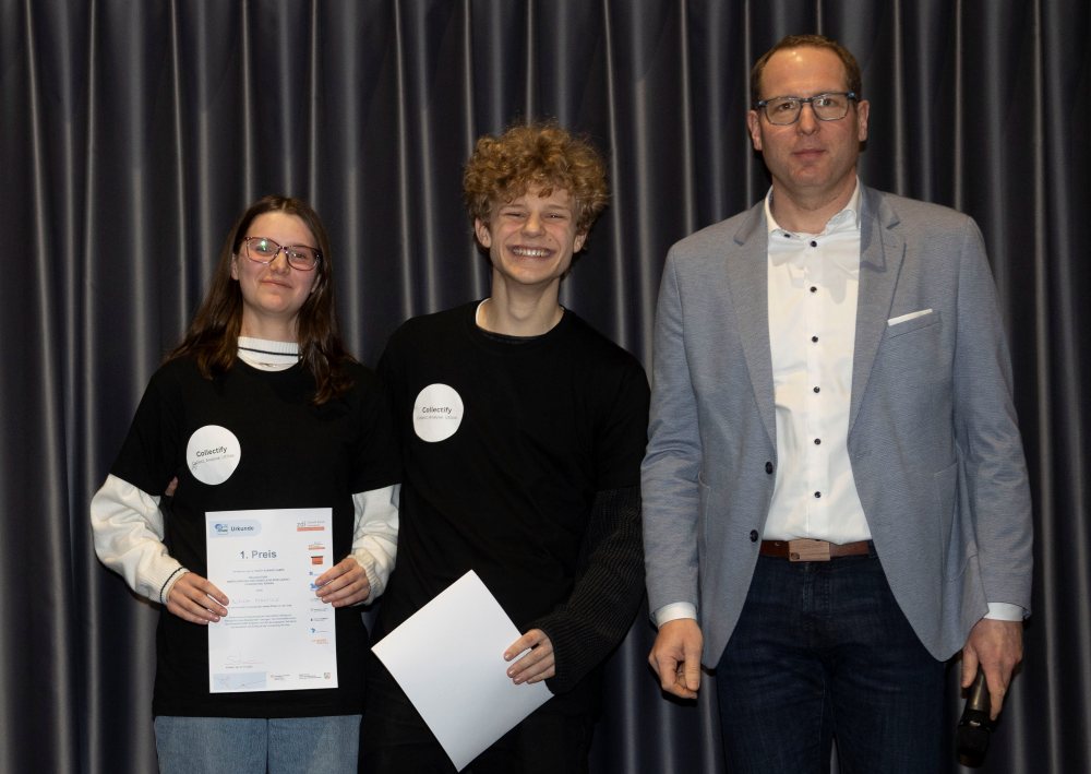 Drei Personen stehen drinnen, zwei halten KReateFUTURE 2025-Zertifikate in den Händen, lächeln und tragen schwarze Hemden mit Namensschildern. Die dritte Person, Dr. Dominik Paul, steht rechts in einem grauen Blazer und mit Brille.