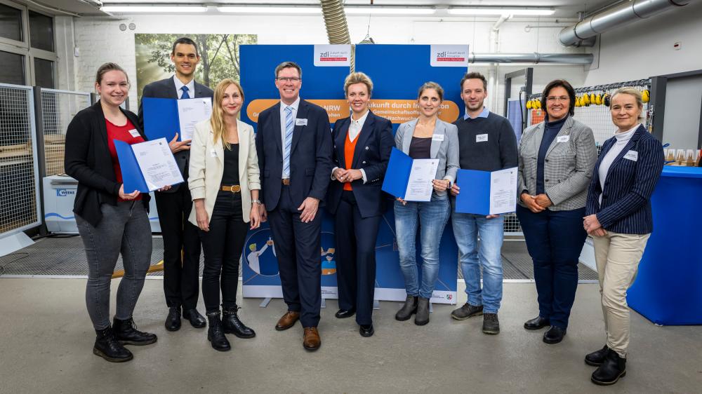 Eine Gruppe von Menschen steht drinnen, hält blaue Mappen mit Förderbescheiden in den Händen und posiert für ein Foto. Sie sind entweder in Business- oder Freizeitkleidung gekleidet, mit einer industriellen Umgebung im Hintergrund.