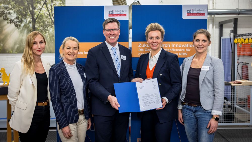 Fünf Personen stehen zusammen, zwei von ihnen halten eine blaue Mappe mit einem Förderbescheid in den Händen. Im Hintergrund sind blaue und orangefarbene Banner zu sehen.