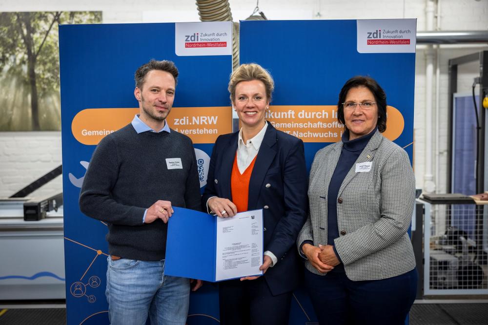 Drei Personen stehen vor einem blauen Hintergrund, die Personen links und in der Mitte halten eine blaue Mappe mit einem Förderbescheid.