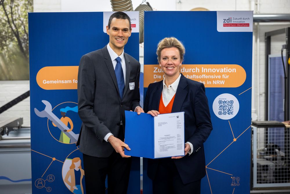 Zwei Personen in formeller Kleidung stehen da, lächeln und halten eine blaue Mappe mit einem Förderbescheid in der Hand. Sie stehen vor einem Hintergrund mit einem zdi-Logo.