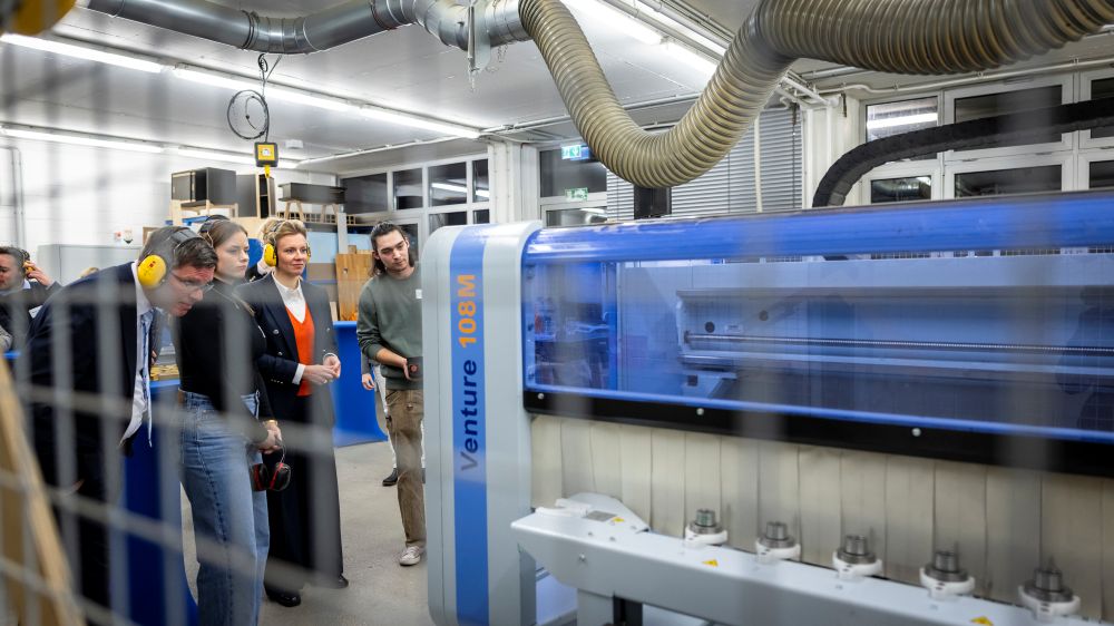 In einer gut beleuchteten Werkstatt beobachtet eine Gruppe von Menschen eine große blaue Industriemaschine. Über ihr verläuft ein komplexes Netzwerk aus Kanälen und Rohren.