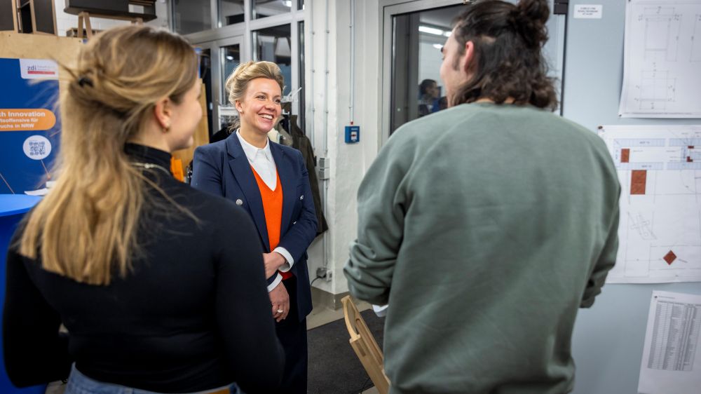 In einer Werkstatt sind drei Personen in ein Gespräch vertieft. Eine Frau im Blazer lächelt, während ein Mann und eine andere Frau ihr zuhören. Im Hintergrund sind die Werkstatt und ein Arbeitsplatz mit Papieren zu erahnen.