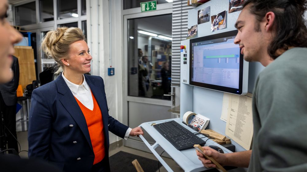 Eine Frau in einem blauen Blazer spricht mit einem Mann, der ein Werkzeug in der Hand hält. Sie befinden sich an einem Arbeitsplatz mit Computerbildschirm in einer Werkstatt. Im Hintergrund ist der Computerbildschirm zu sehen, auf dem Diagramme angezeigt werden.