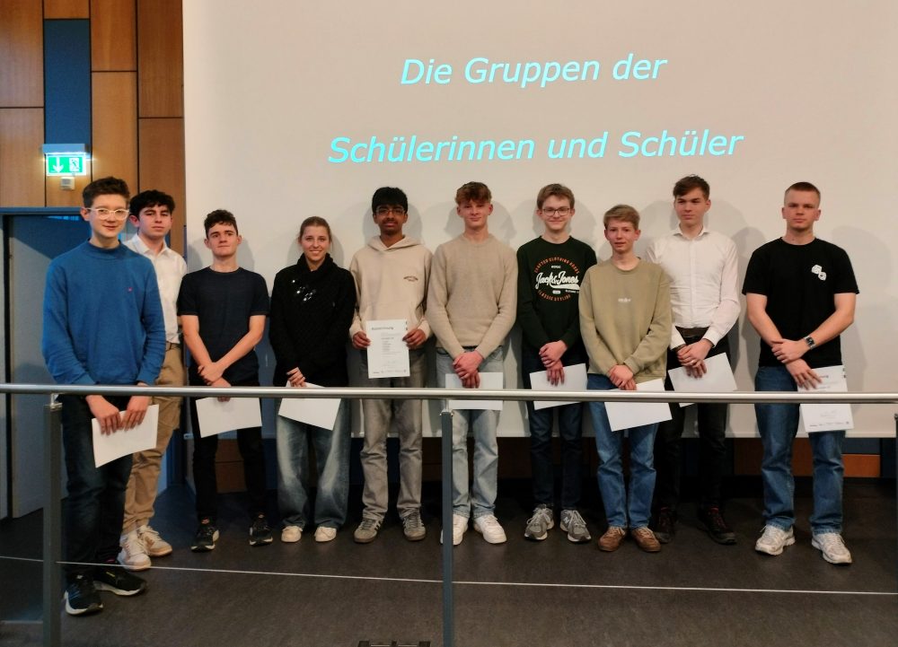 Zehn Schüler:innen stehen vor einem Bildschirm auf dem steht "Die Gruppen der Schülerinnen und Schüler". Sie halten jeweils ein Blatt Papier in der Hand und posieren versetzt in einer Reihe.
