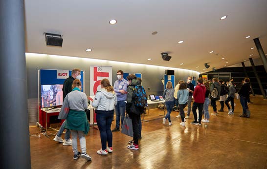 In einer modernen Ausstellungshalle versammeln sich Menschen zwischen verschiedenen Ständen und Ausstellungen und kommen mit den Ausstellern ins Gespräch.