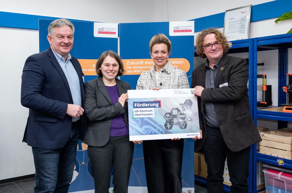 Vier Personen stehen drinnen und halten ein großes Schild hoch, auf dem sie über die Finanzierung des zdi-Zentrums Oberhausen sprechen, einer wichtigen MINT-Initiative im Rahmen der Netzwerkförderung. Der Hintergrund ist in Blau- und Orangetönen gehalten.