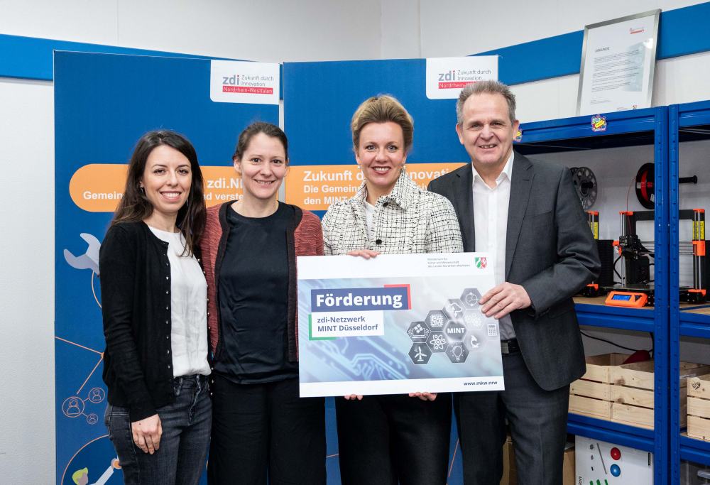 In einer Büroumgebung halten vier Personen stolz ein großes Schild mit dem Bezug zum zdi-Netzwerk MINT Düsseldorf. Im Hintergrund betonen Regale und ein Plakat mit Text ihr Engagement für die Netzwerkförderung.