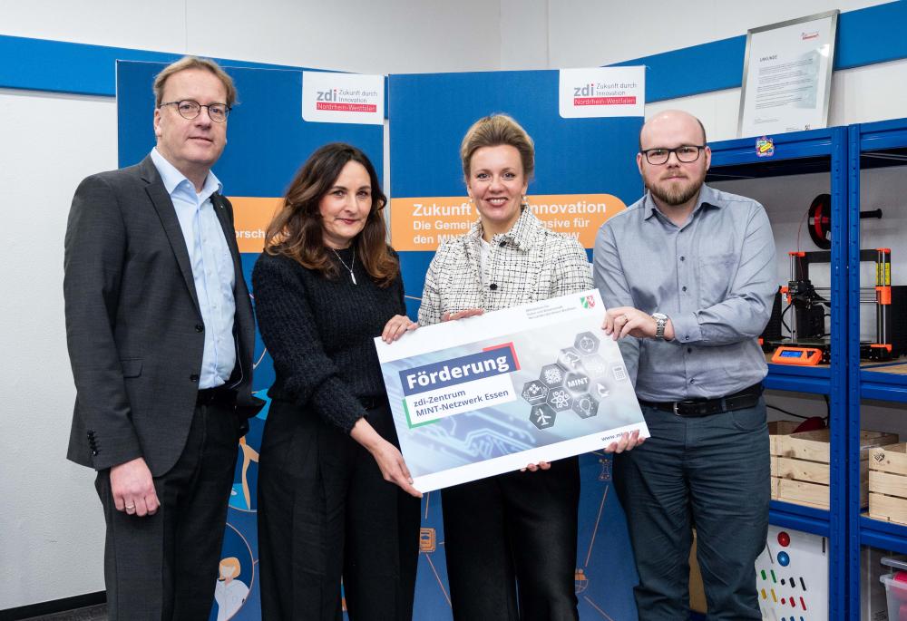Vier Personen stehen in einem Büro, zwei von ihnen halten ein großes Plakat mit der Aufschrift „Förderung“ in den Händen, das ihr Engagement für die Netzwerkförderung symbolisiert. Im Hintergrund arbeitet ein 3D-Drucker, der der Szene einen innovativen Touch verleiht.