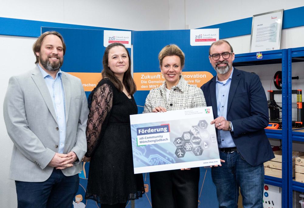 Vier Personen stehen in einem Raum und halten ein großes Schild mit einem Text zur Förderung. Im Raum stehen blaue Regale und Poster einer deutschen Innovationsinitiative. Diese Elemente unterstreichen die Wirkung der Netzwerkförderung bei der Förderung der Zusammenarbeit und des technologischen Fortschritts.