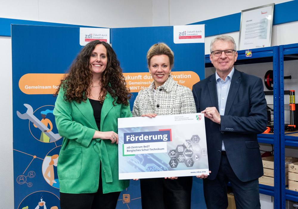 Drei Personen stehen stolz in einem Raum mit Bannern und Equipment und halten ein großes Schild hoch, das ihr Engagement für die Netzwerkförderung betont.