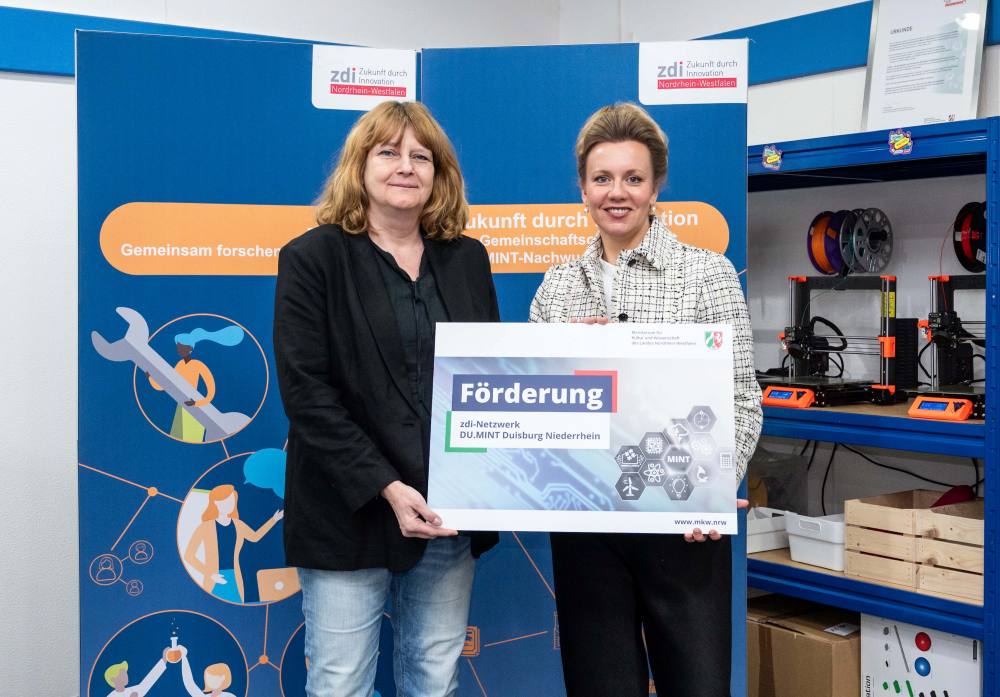 Zwei Personen stehen vor einem technisch gestalteten Hintergrund und halten ein Schild mit der Aufschrift „Förderung“. In der Kulisse sind Regale mit 3D-Druckern zu sehen, die das Konzept der Netzwerkförderung hervorheben.