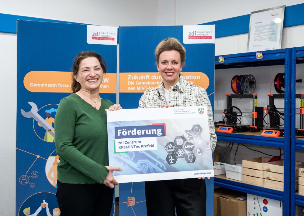 Zwei Menschen stehen lächelnd mit einem Schild in der Hand in einem Raum voller Plakate und Regale mit diversem Equipment. Sie sind begeistert von der Netzwerkförderung.