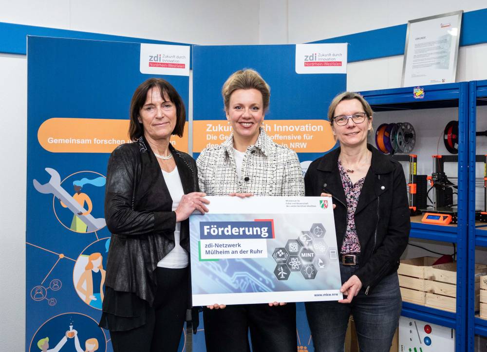 Drei Frauen stehen in einem Technik-Themenraum zusammen und halten ein Schild mit der Aufschrift „Förderprogramm“. Im Hintergrund sind Transparente mit der Aufschrift „Netzwerkförderung“ zu sehen.