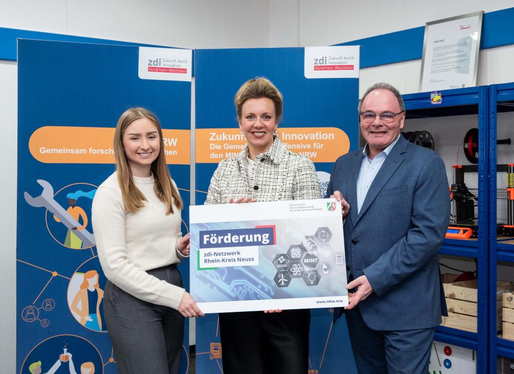 Drei Menschen halten stolz ein Schild der „Förderung zdi-Netzwerk Rhein-Kreis Neuss“ in die Höhe, stehen vor Bannern mit dem zdi-Logo und feiern die erfolgreiche Netzwerkförderung.