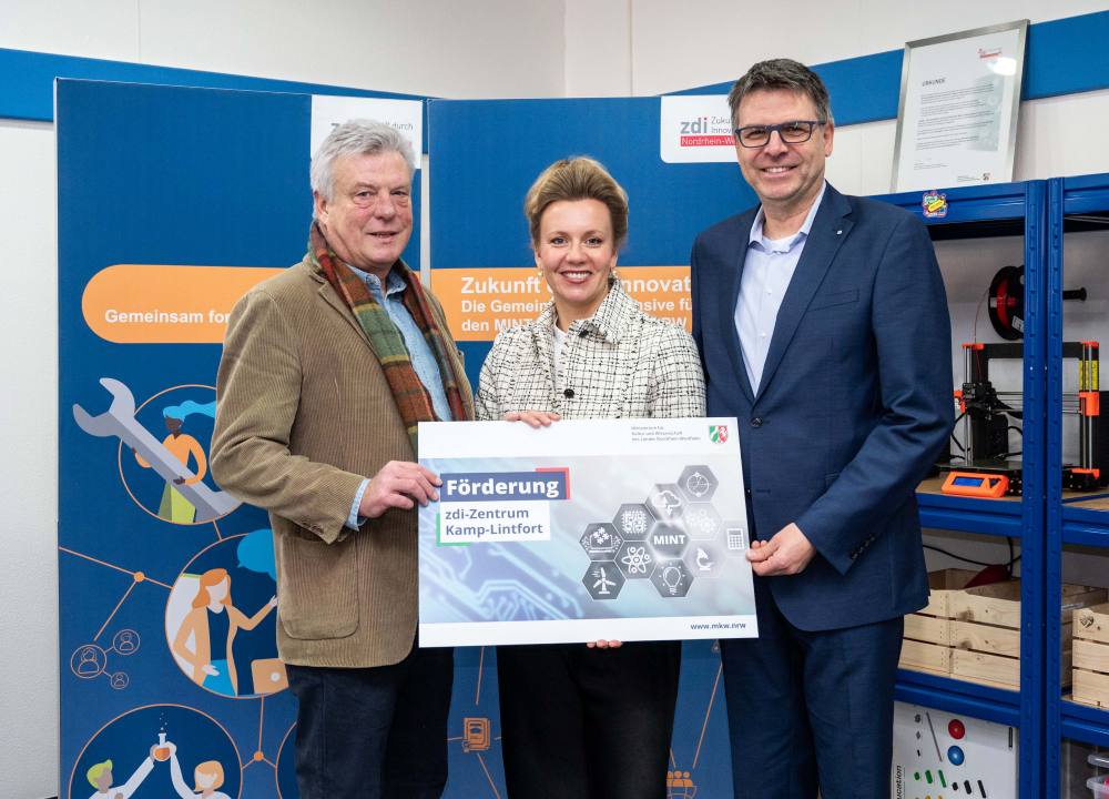 Drei Personen stehen im Innenbereich und halten stolz ein Schild mit der Netzwerkförderung in die Höhe. Dahinter bildet eine Kulisse mit Industriesymbolen die Kulisse, und Regale voller Ausrüstung lassen auf die vielversprechenden Projekte schließen, die durch ihre Förderung unterstützt werden.