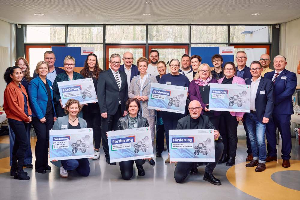 Eine Gruppe von Menschen in steht im Innenbereich einer Schule, lächelt in die Kamera und hält große Schilder in den Händen.