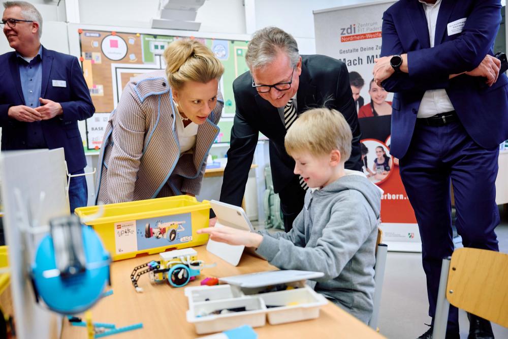 In einem Klassenzimmer zeigt ein Kind mehreren Erwachsenen etwas an einem Tablet, während Unterrichtsmaterialien auf dem Tisch liegen.